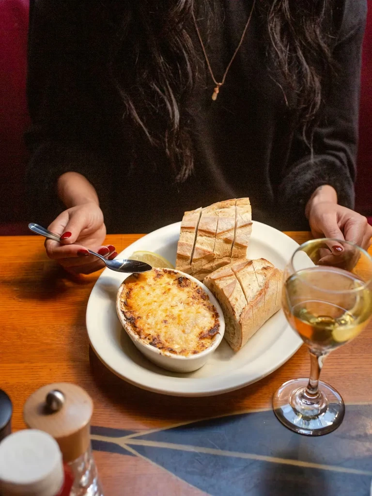 Close-up of gratin, baguette, wine, and hand with spoon on table.