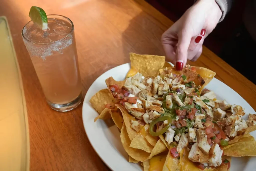 A hand reaches for loaded chicken nachos with a pink cocktail.
