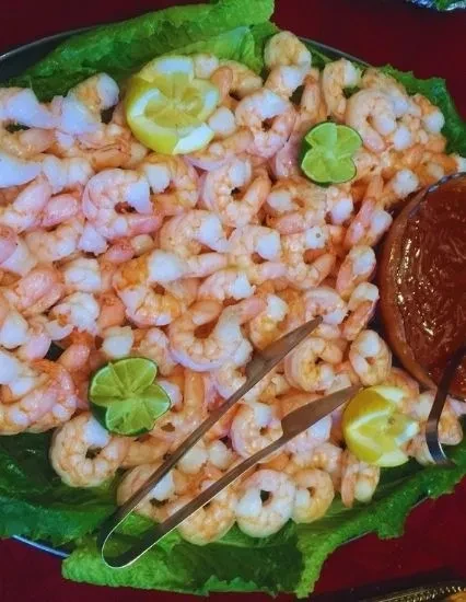 Platter of peeled shrimp with lettuce, lemon, lime, cocktail sauce, tongs.