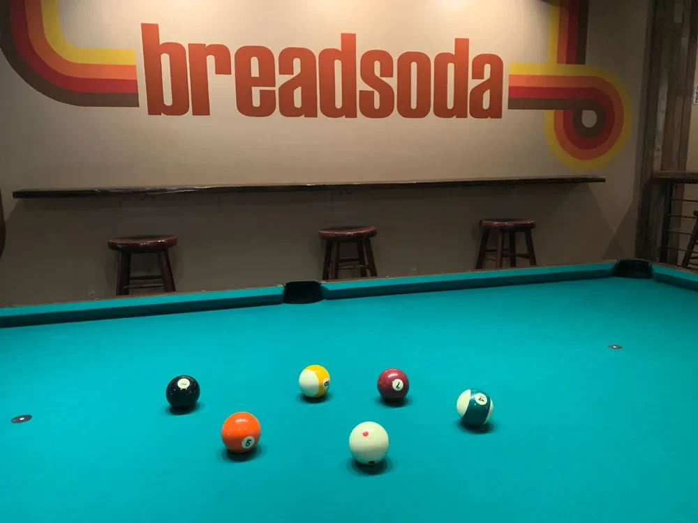 Turquoise pool table with billiard balls and a retro "breadsoda" wall graphic.