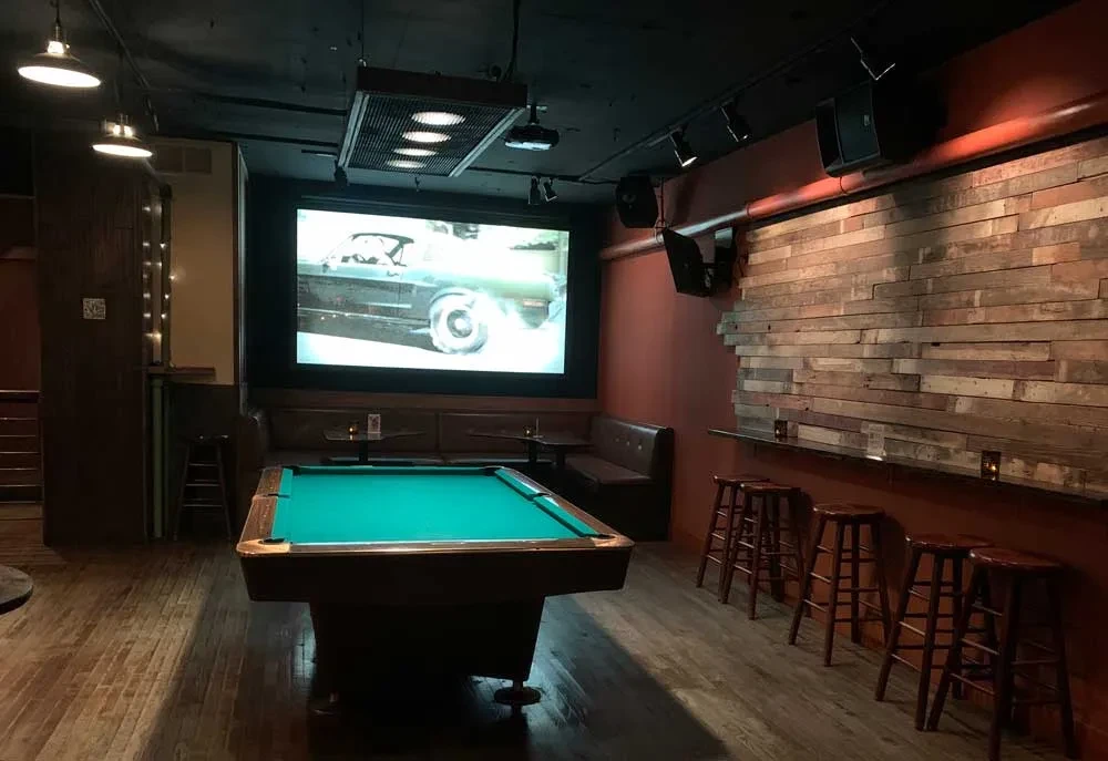 Dimly lit industrial bar with pool table and vintage car on screen.