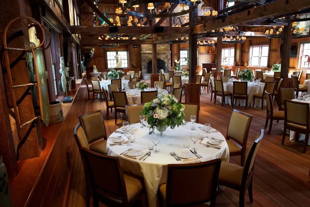 Elegant rustic dining hall with wood beams, set tables, and fireplace.