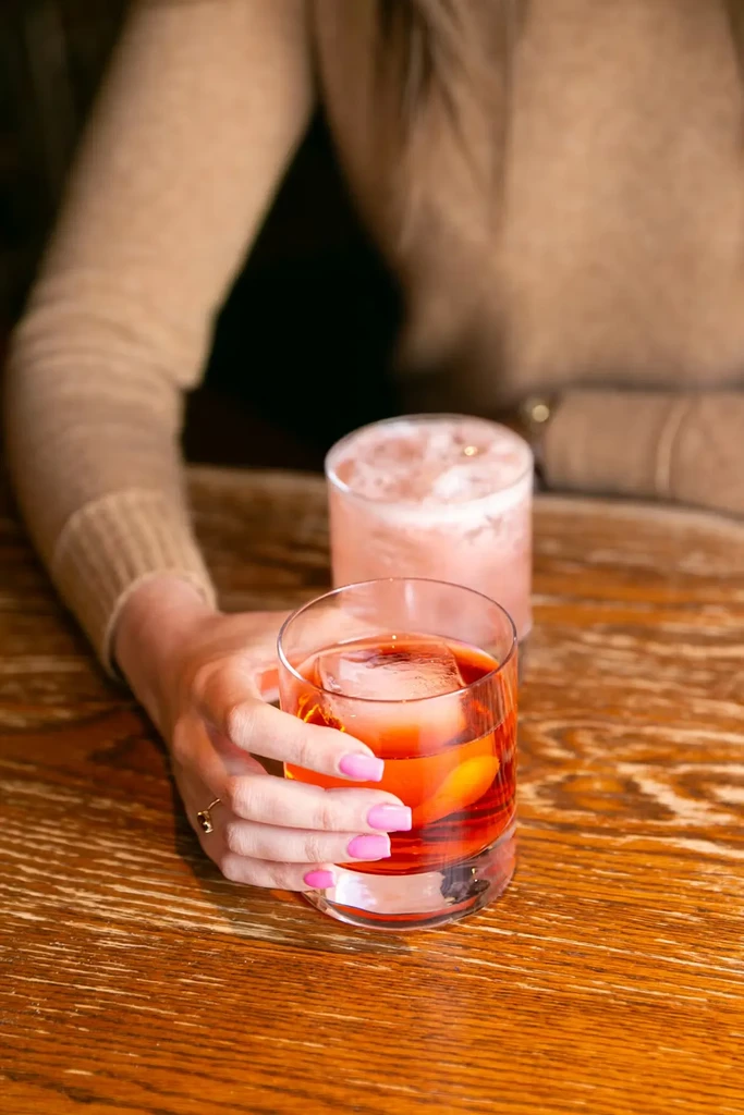 Pink-nailed hand holds amber cocktail; another blurred drink on wood table.