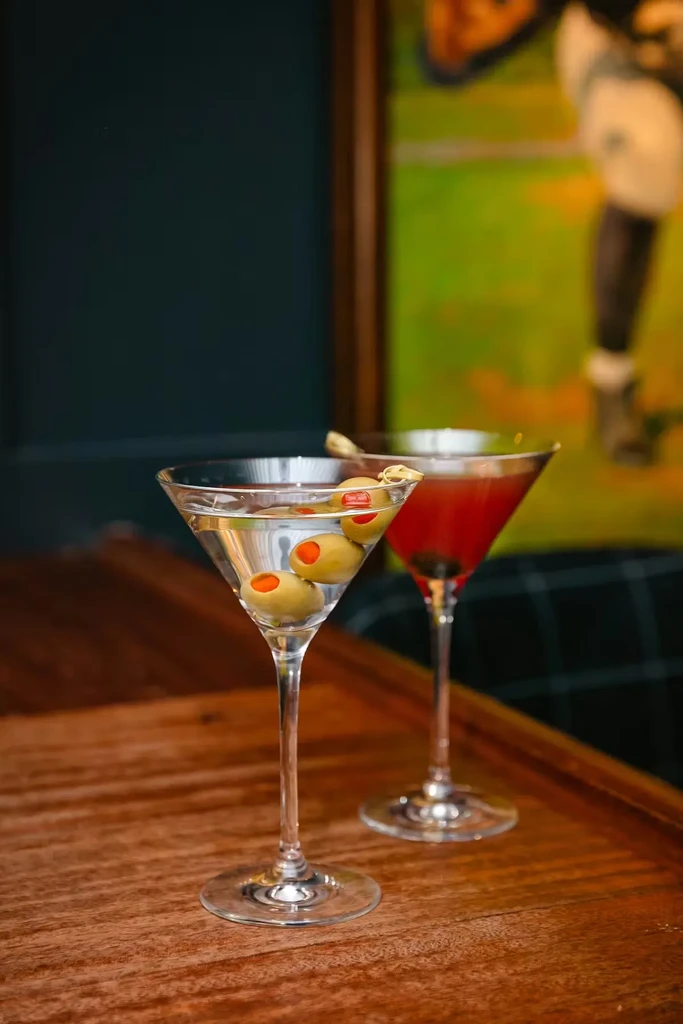 Martini with olives and red cocktail on a polished wood surface.