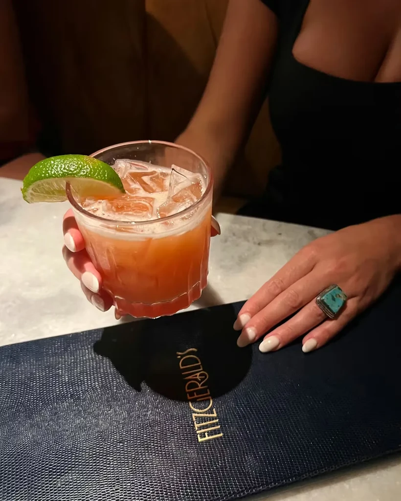 Orange cocktail with lime held over dark blue Fitzgerald's menu.
