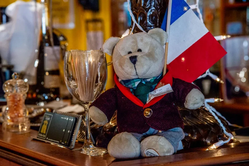 Plush teddy bear in red jacket holding a French flag on a bar.