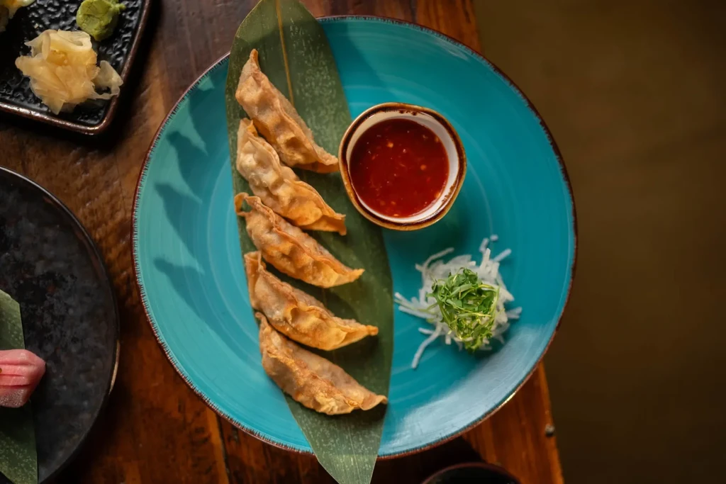 Five pan-fried dumplings on green leaf, turquoise plate, red sauce.