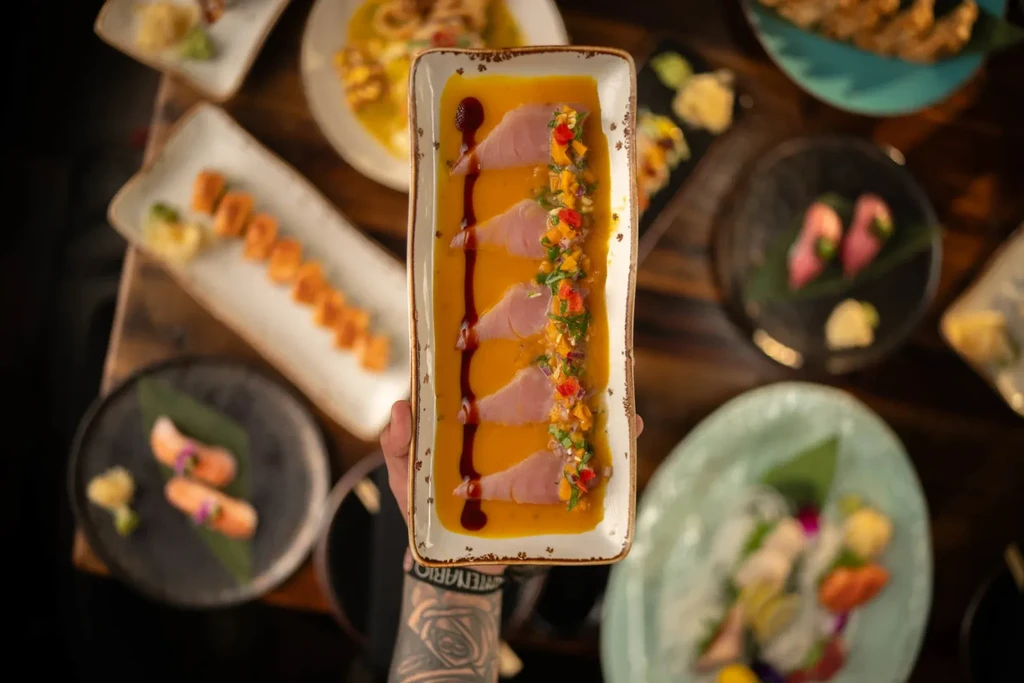 Tattooed hand holding a gourmet sashimi dish with colorful garnishes.