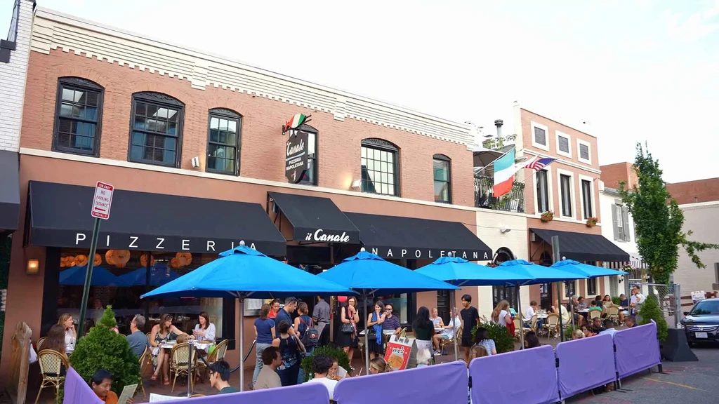 Exterior of il Canale Pizzeria, with lively outdoor dining and blue umbrellas.