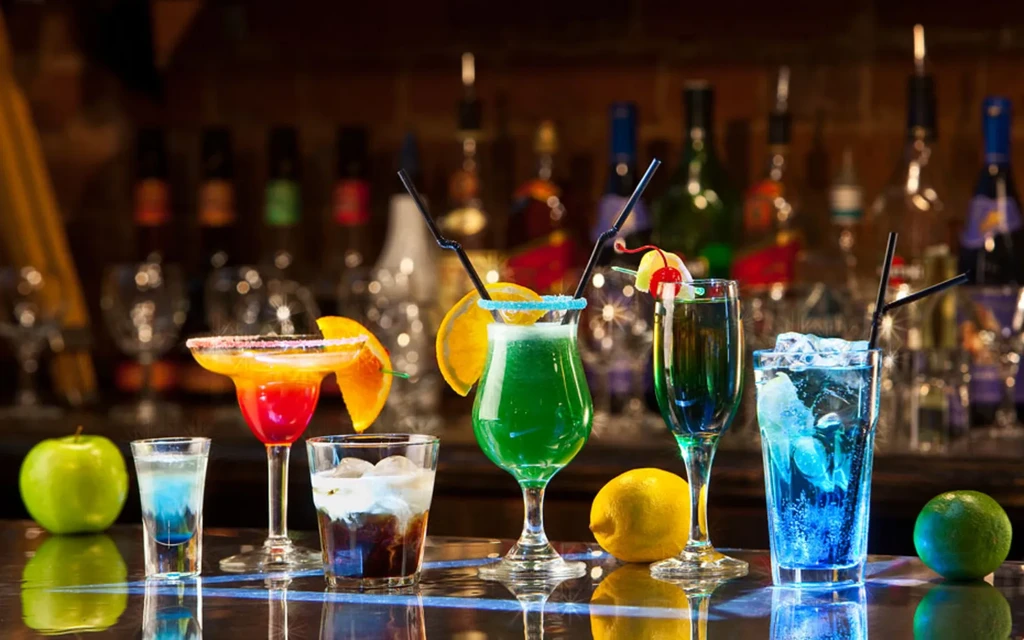 Seven colorful cocktails and shots on a bar counter with fruits.