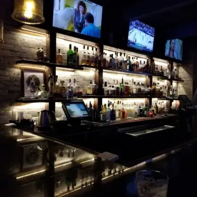 Dimly lit bar with illuminated liquor wall and three flat-screen TVs.
