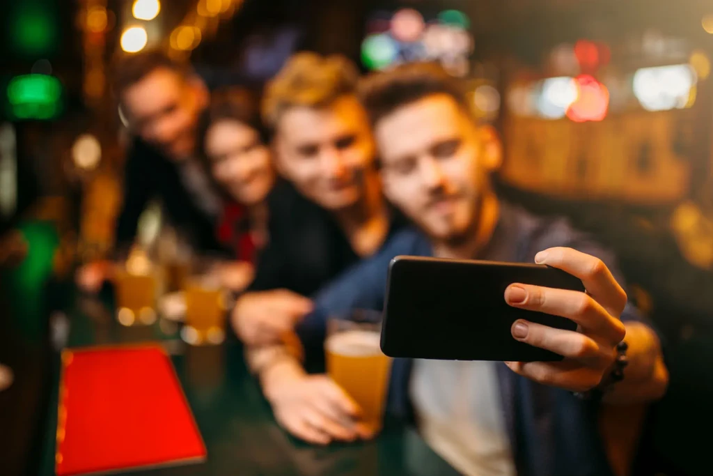 Blurred selfie of four people with drinks in a busy bar.