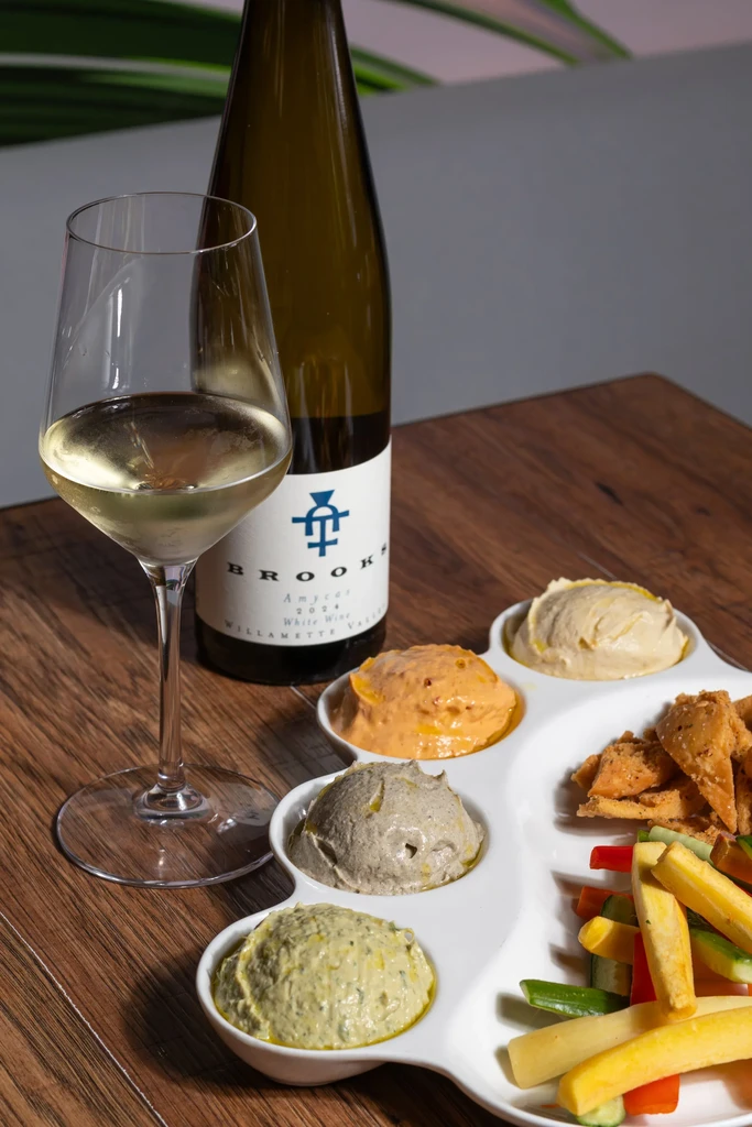 White wine bottle, glass, dips, pita chips, vegetables on wooden table.