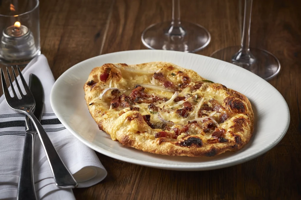 Small pizza with cheese, bacon, onions, cutlery, wine glasses, and candle.