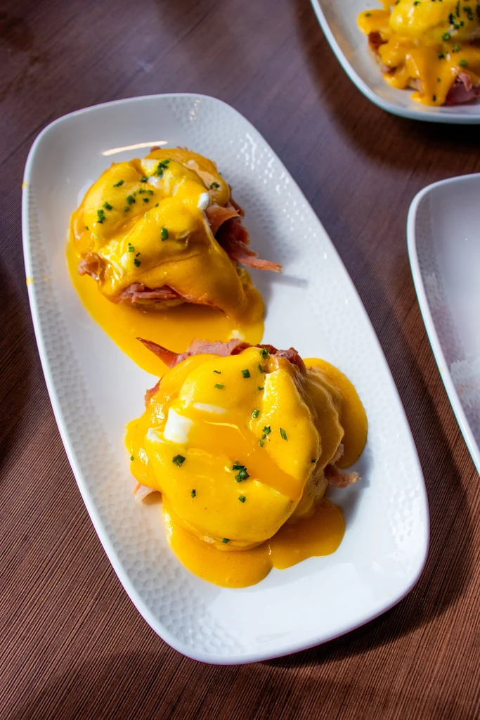 Two plates of Eggs Benedict with runny yolks, hollandaise, and green herbs.