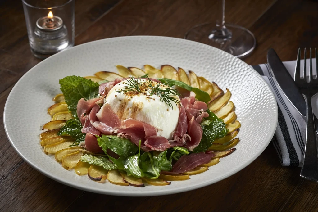 Plated burrata with prosciutto, greens, and potato wedges in a restaurant.
