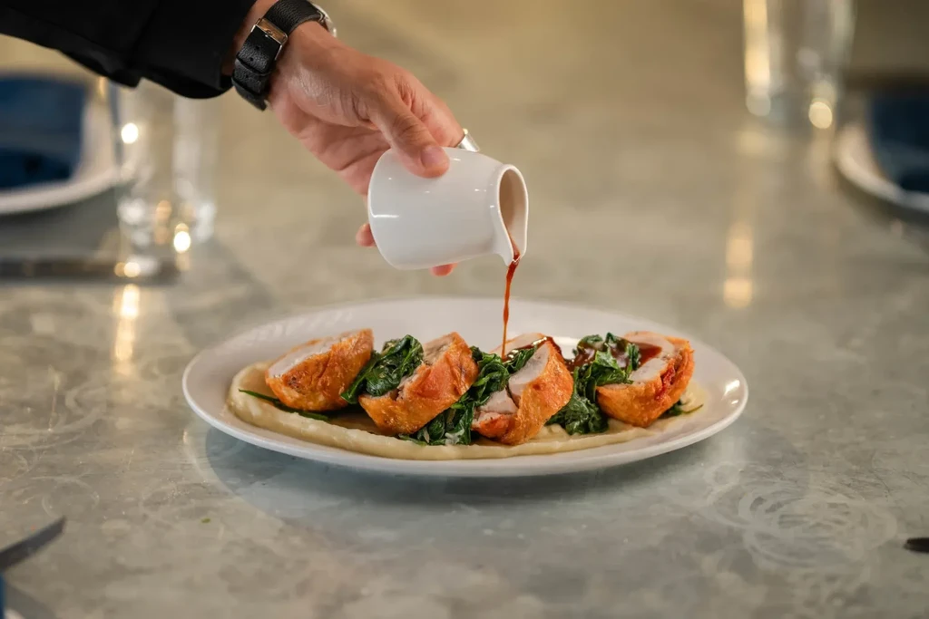 A hand pours red sauce over five chicken segments and greens.