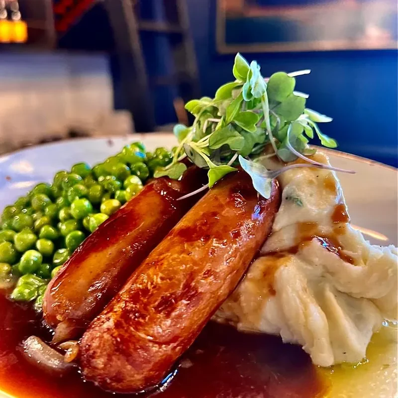 Sausages, mashed potatoes, peas, and gravy, garnished with microgreens.