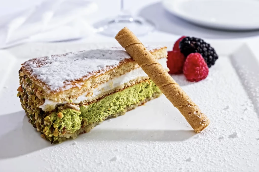 Plated multi-layered dessert with green layer, nuts, berries, and wafer.