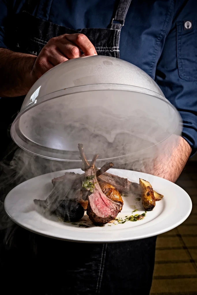 Hands lift cloche from medium-rare rack of lamb releasing white smoke.