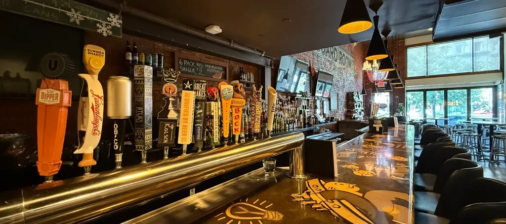 Modern bar interior with long counter, numerous beer taps, and brick walls.