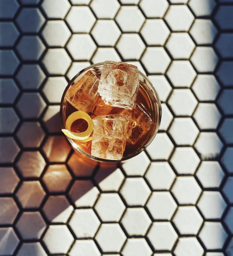 Top-down: Amber drink with ice and lemon peel on hexagonal tiles.