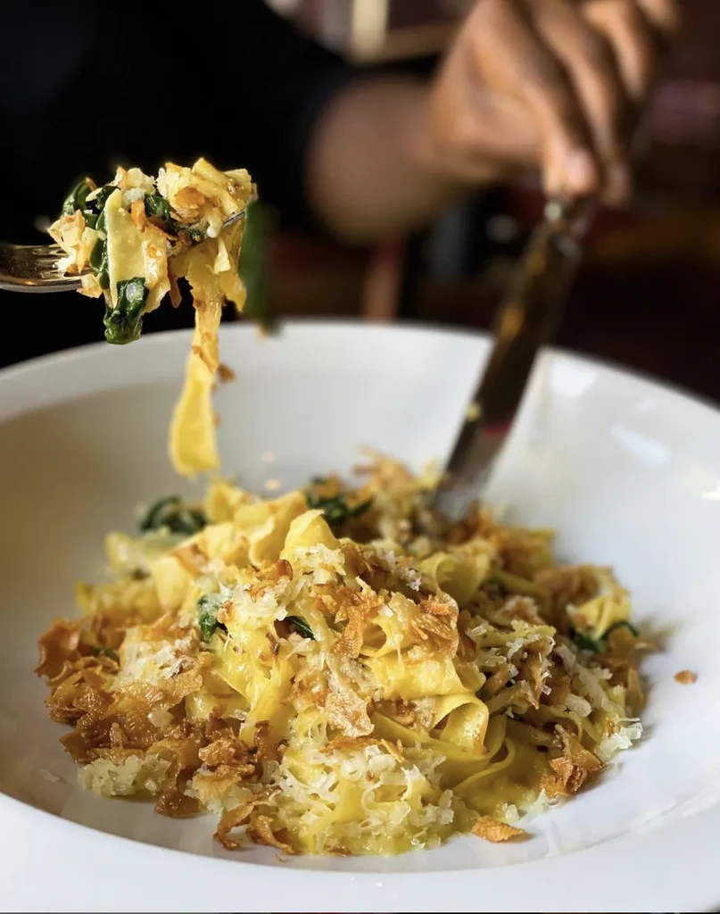 A hand lifts cheesy fettuccine pasta with greens and crispy topping.