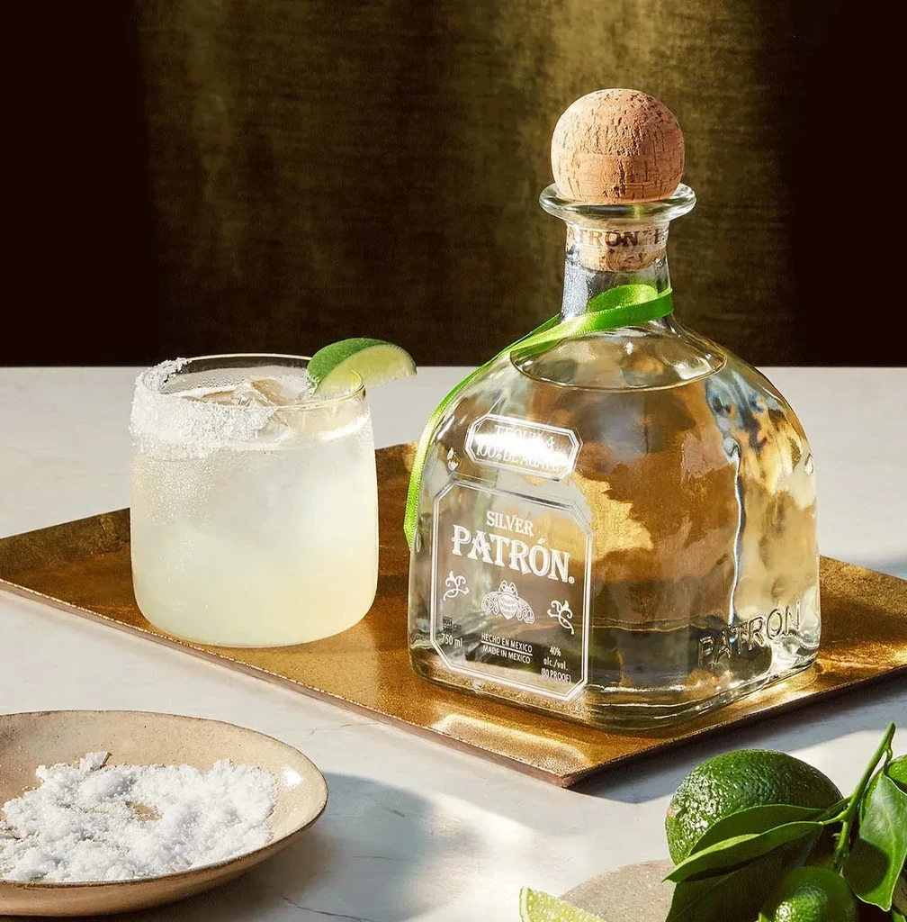 Patrón Silver tequila bottle, cocktail, limes, and salt on golden tray.
