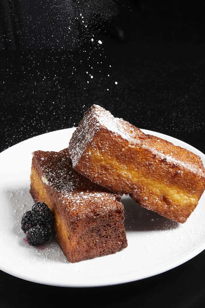 Stacked French toast, powdered sugar being sprinkled, with blackberries.