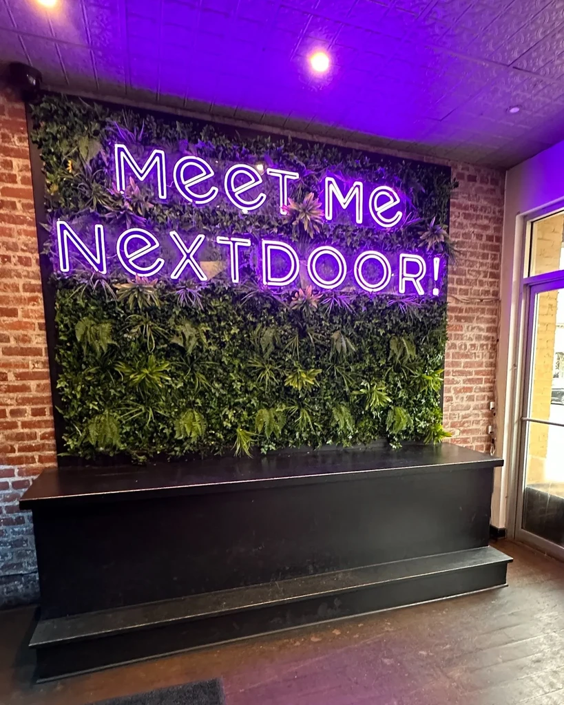 Faux plant wall with "Meet Me NEXTDOOR!" neon sign, purple lighting.