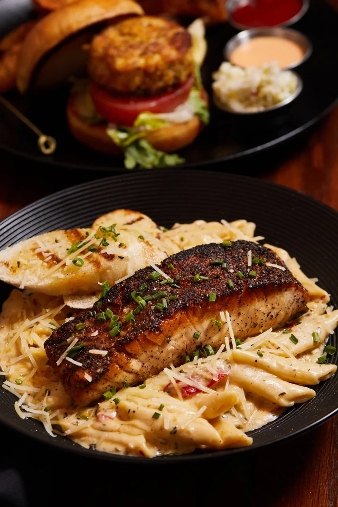 Close-up of penne pasta with seared fish, plus blurred burger and sides.
