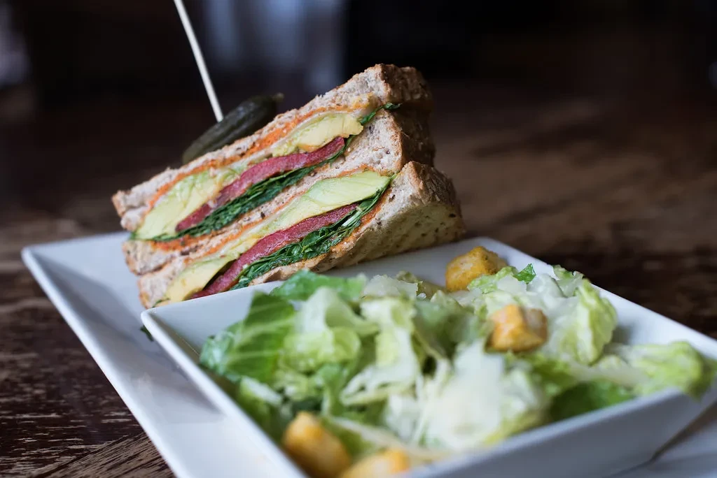 Sliced whole-grain sandwich with avocado, peppers, greens, and a side salad.