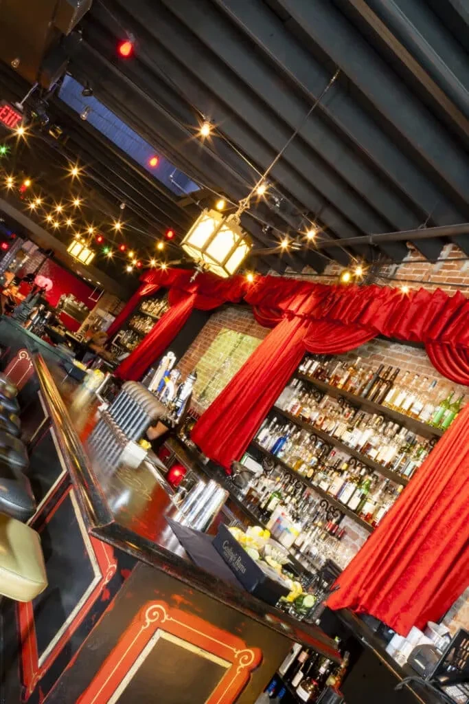 Dramatic bar with ornate dark wood counter, illuminated liquor, and velvet drapes.