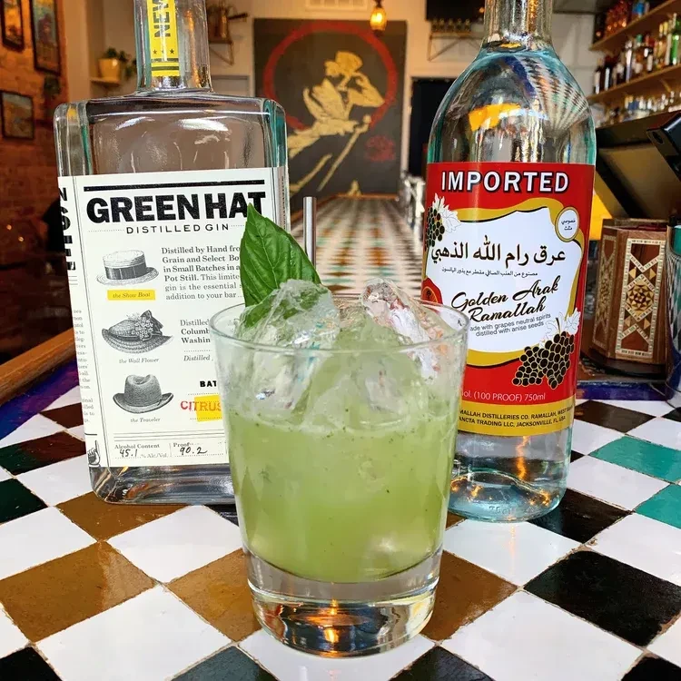 Green cocktail, ice, and two liquor bottles on a checkered bar.