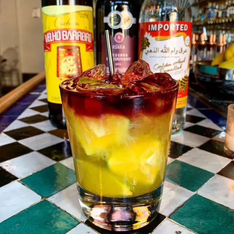 Vibrant layered cocktail with reddish ice and metal straw on checkerboard.