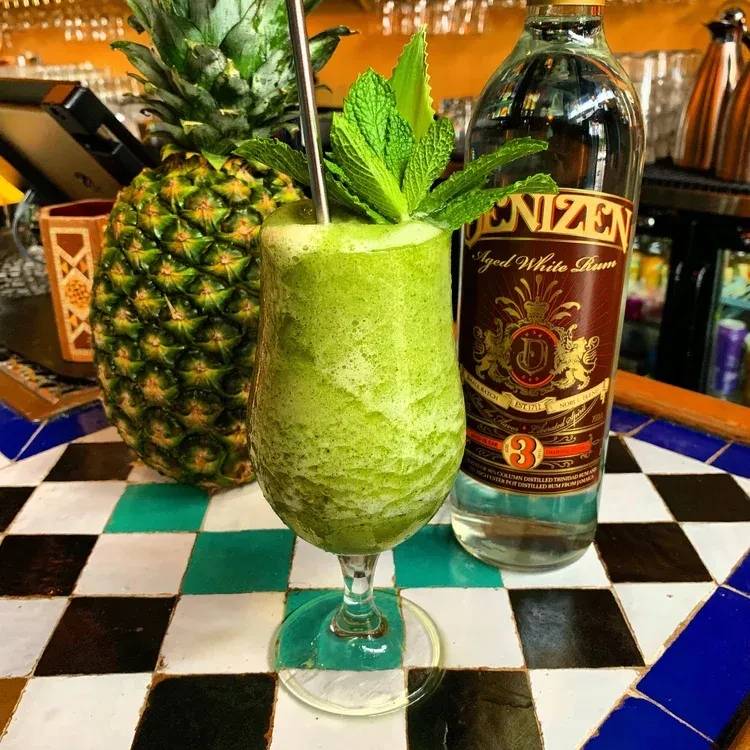 Green drink, pineapple, and Denizen rum bottle on checkered tiles.