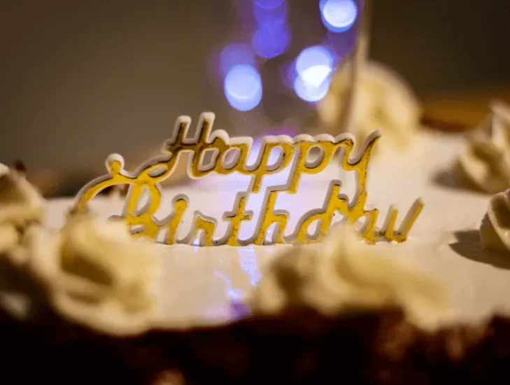 Gold "Happy Birthday" topper on a frosted cake with blurred lights.