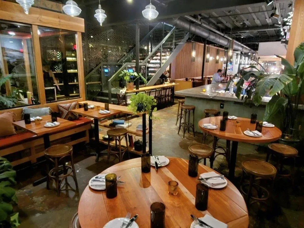 Modern restaurant interior with wooden tables, bar, plants, and a person.