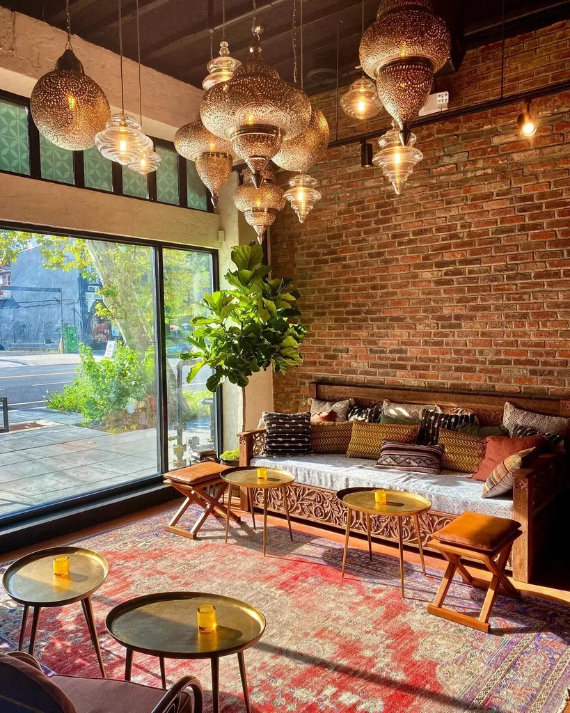 Eclectic lounge with Moroccan chandeliers, cushioned bench, and exposed brick wall.