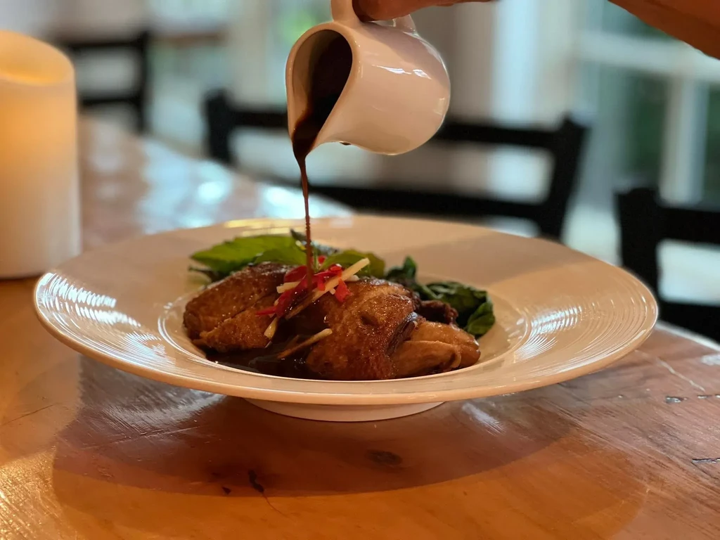Dark sauce being poured onto plated poultry and vegetables.