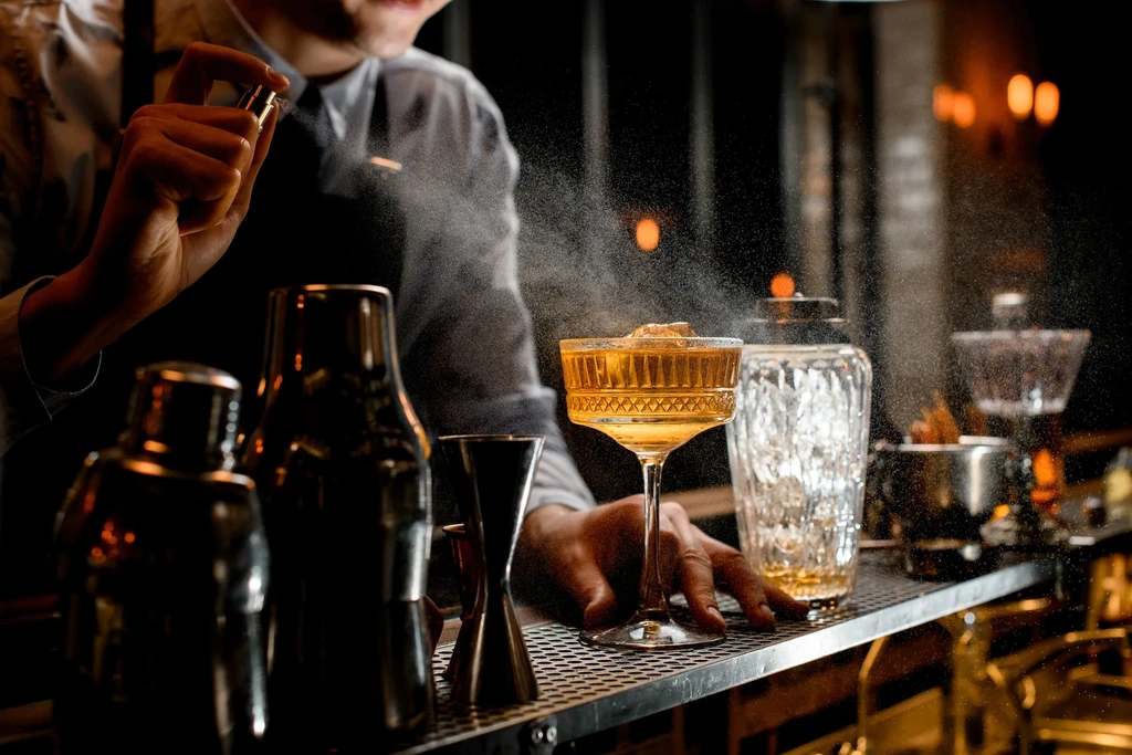 Bartender mists a golden cocktail in an ornate stemmed glass.
