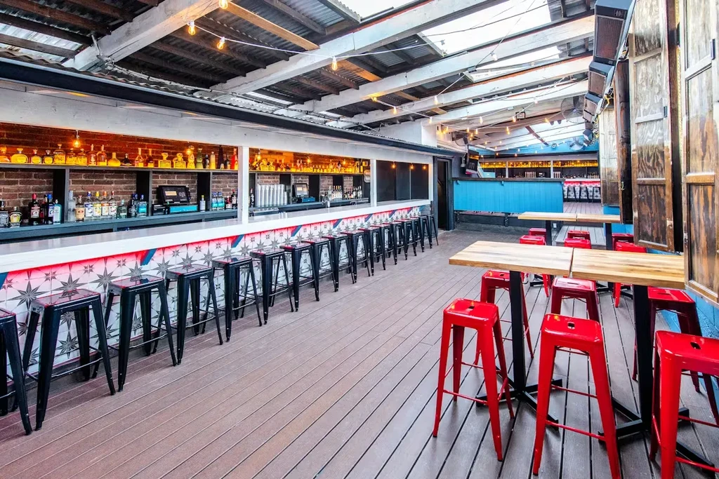 Open-air bar with colorful tiled counter, spirit bottles, and red high-top stools.