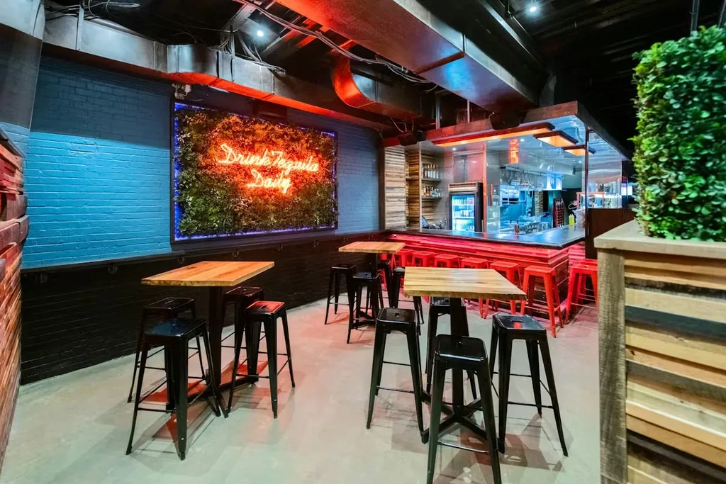 Industrial-chic bar with 'Drink Tequila Daily' neon sign on plant wall.