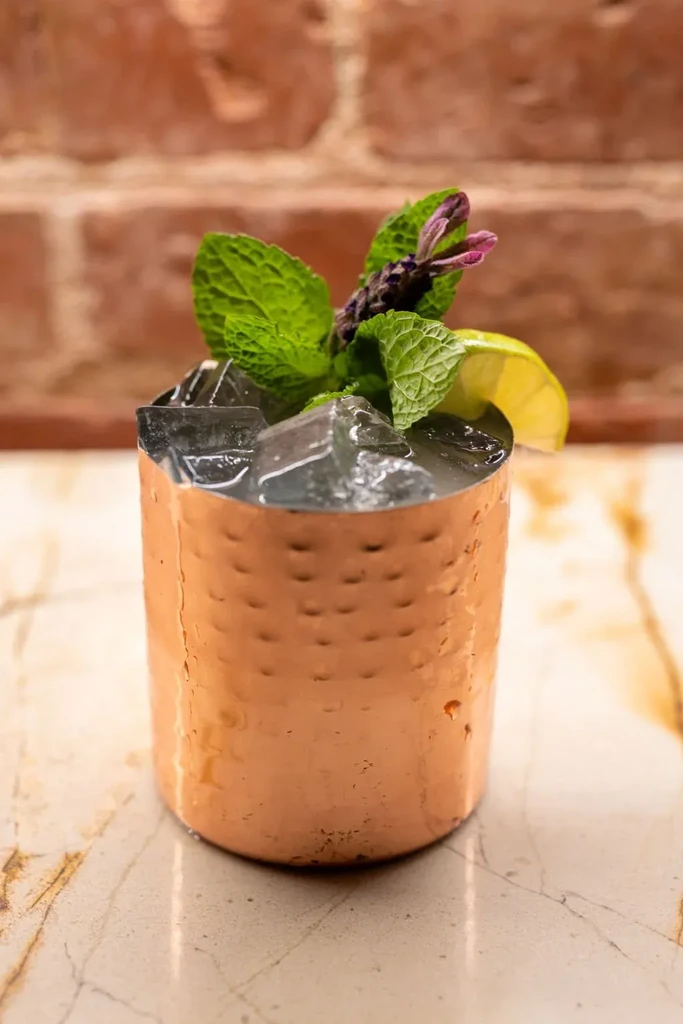 Hammered copper mug cocktail with mint, purple sprig, lime on counter.