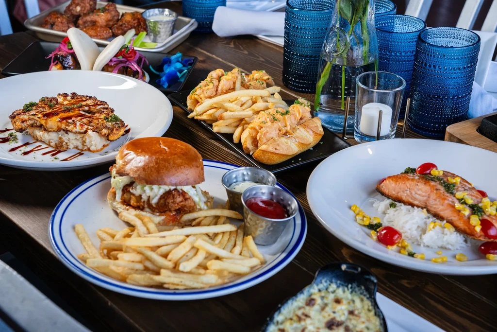Dark wooden table with burger, salmon, bao buns, fries, and drinks.