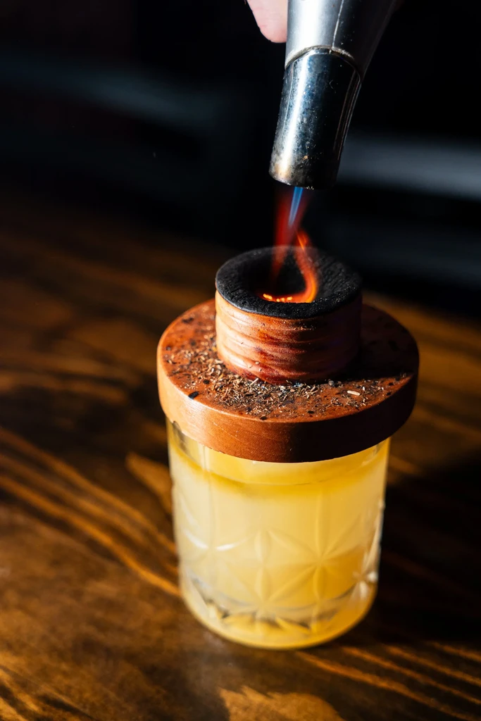 A hand torches wood chips to smoke a cocktail.
