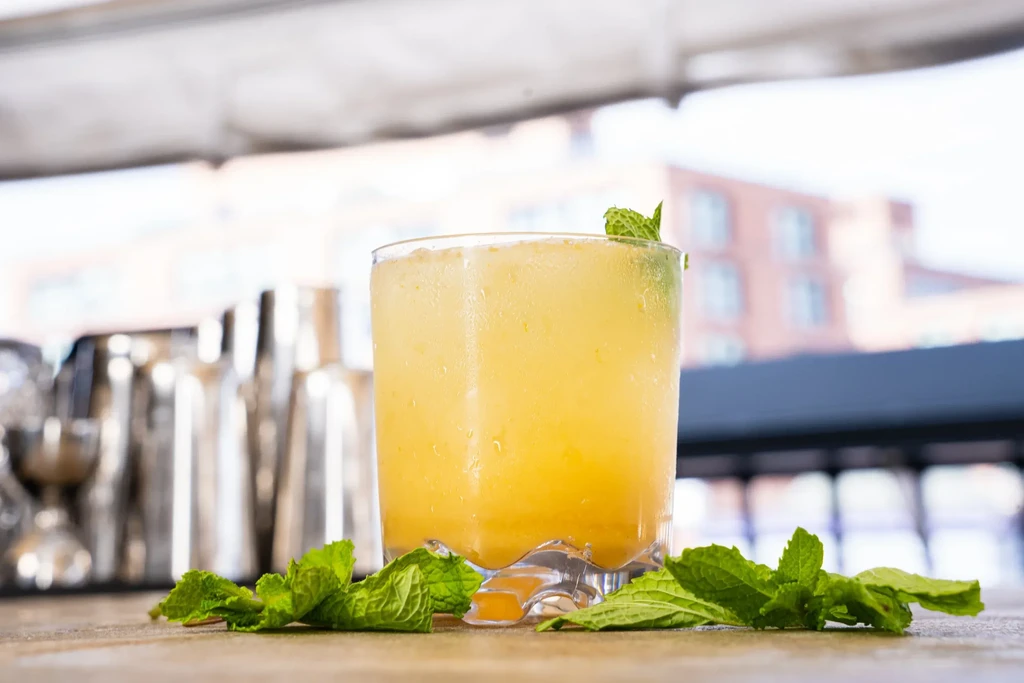 Chilled yellow-orange cocktail with mint in faceted glass on a bar.