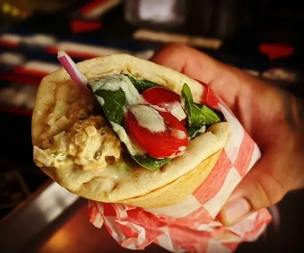 Hand holds a pita wrap with creamy filling, vegetables, and tomatoes.