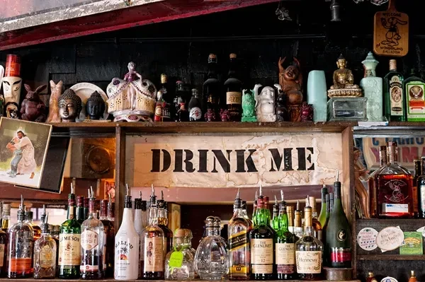 Eclectic bar shelf with liquor bottles and a "DRINK ME" sign.