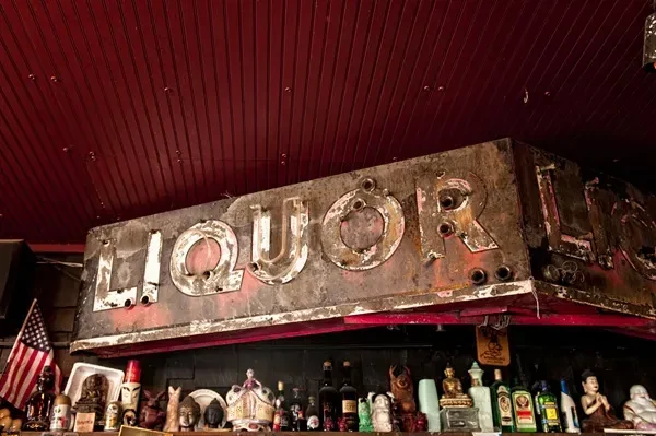 Rusty LIQUOR sign above a shelf of bottles, Buddha figures, flag.